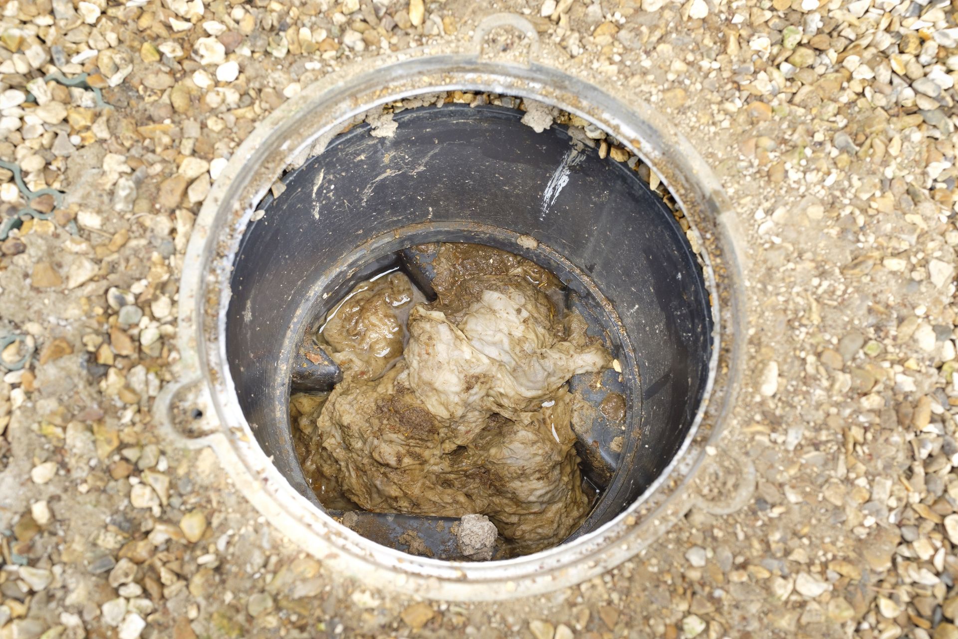 Open manhole with a mass of brown, fibrous debris inside, set in gravel.