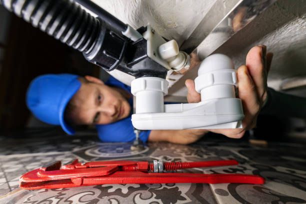 Plumber in blue cap repairs pipes under sink, using a wrench on a tiled floor.