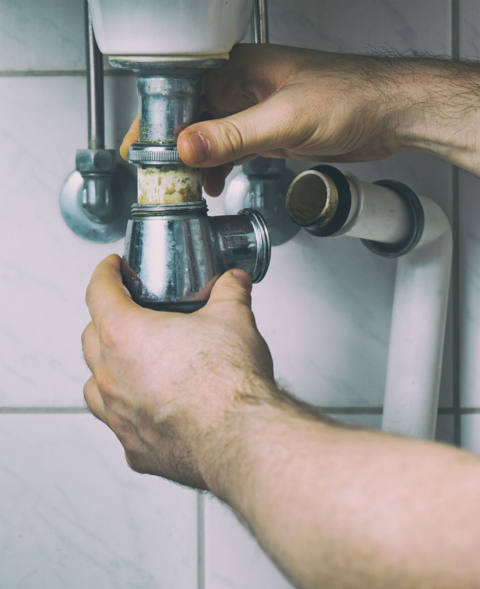 Person's hands assembling plumbing under a sink; chrome pipes against white tile.