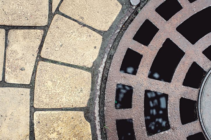 Circular metal manhole cover with square cutouts next to light-colored, curved brick pavement.