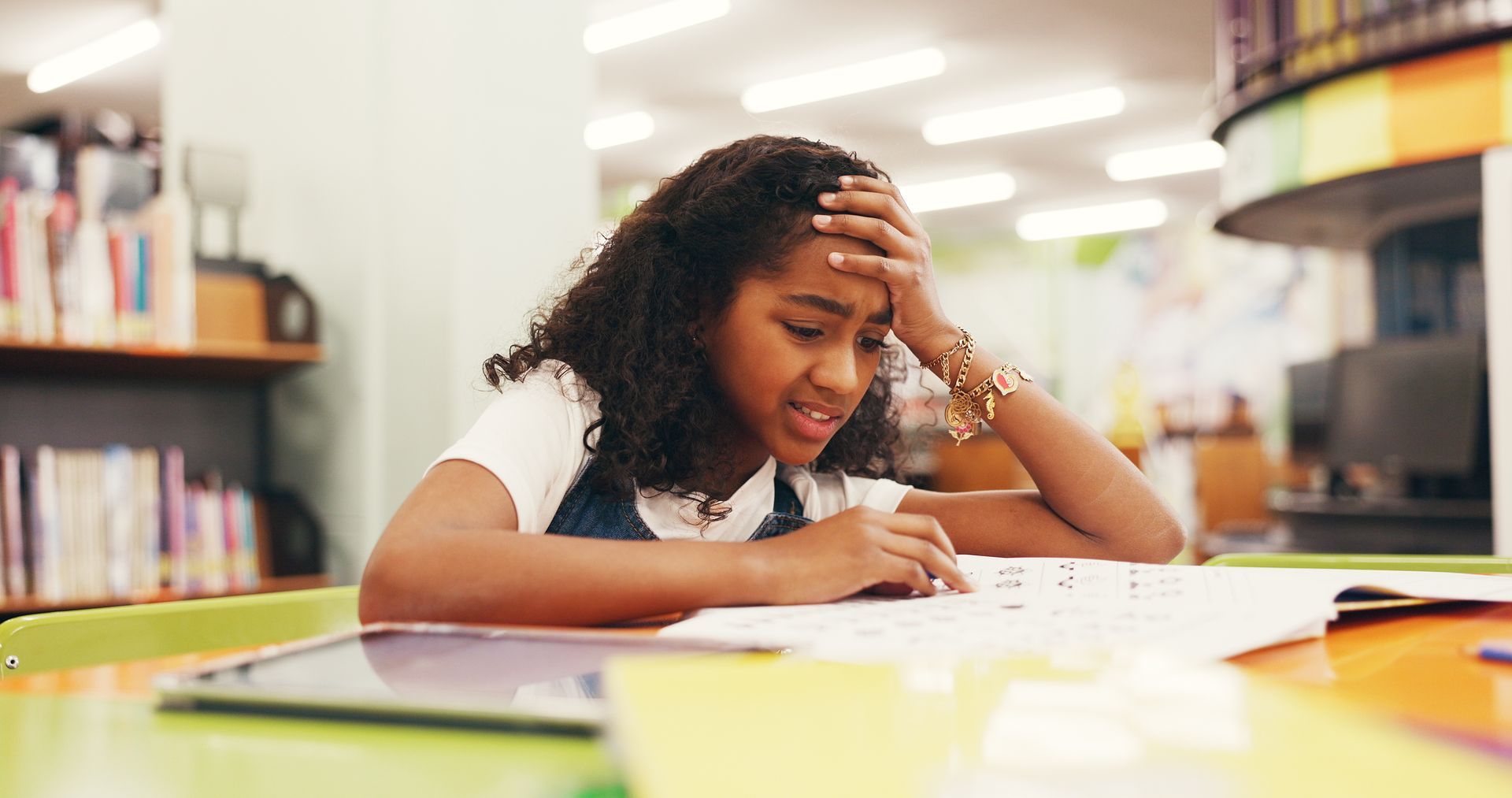 Student studying at a library table, reading a book with a frustrated expression.