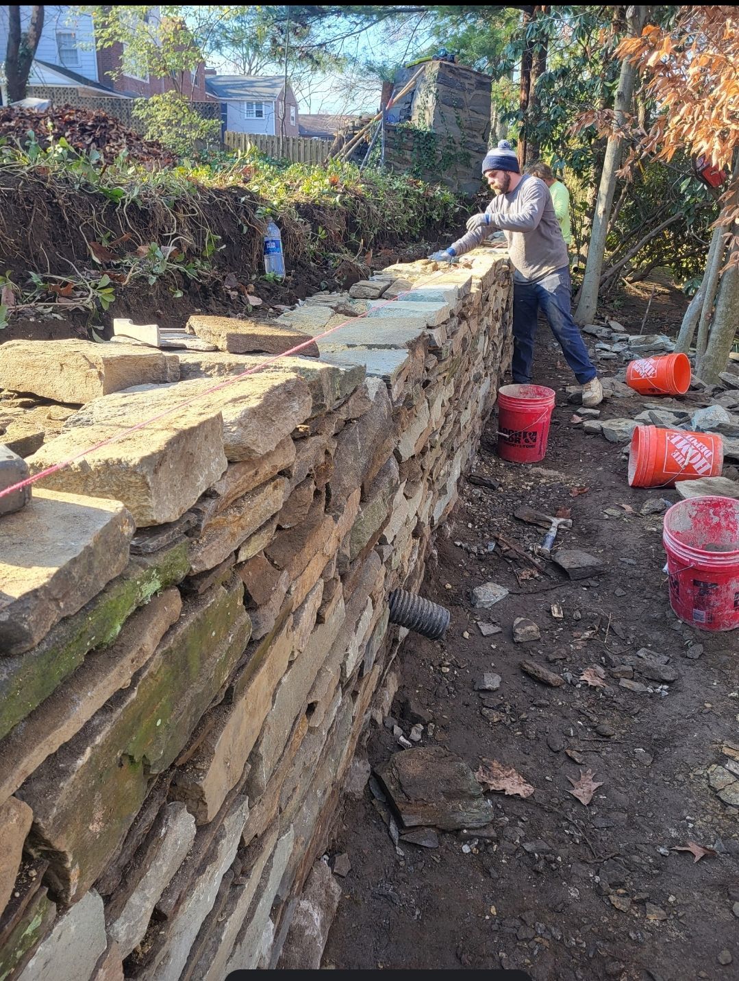 Person building a stone wall outdoors; setting stones with mortar, using red buckets.