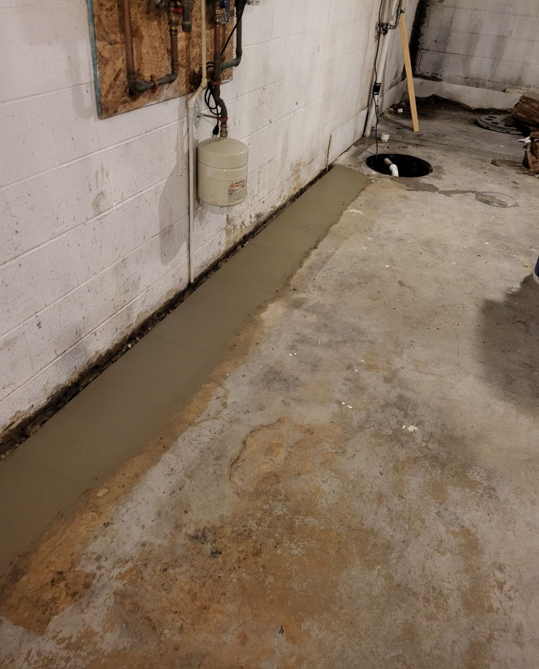 Basement with concrete floor, newly poured cement along wall, pipes, and utility tank.