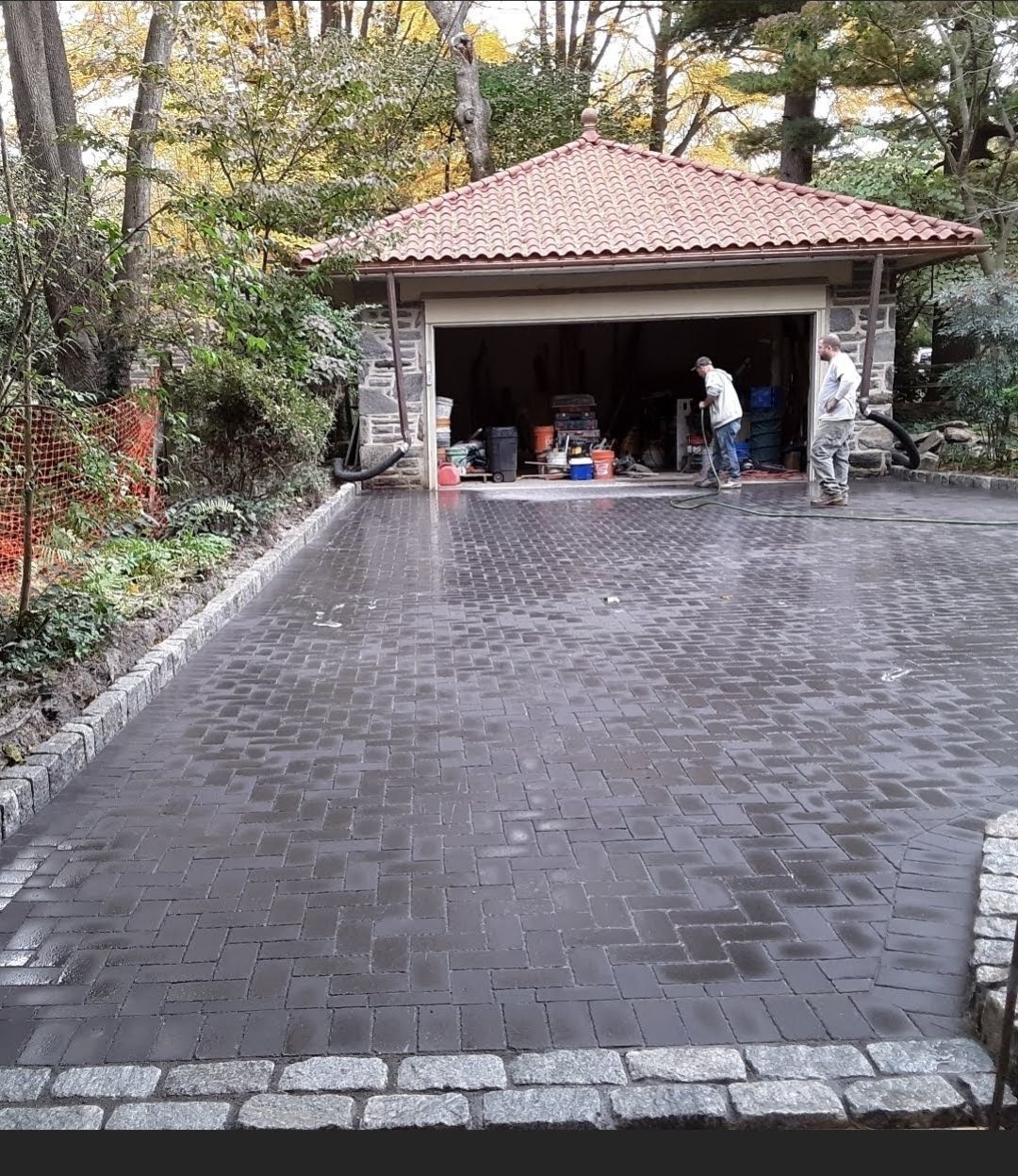 Brick driveway leading to a garage with two people working inside. Trees surround.