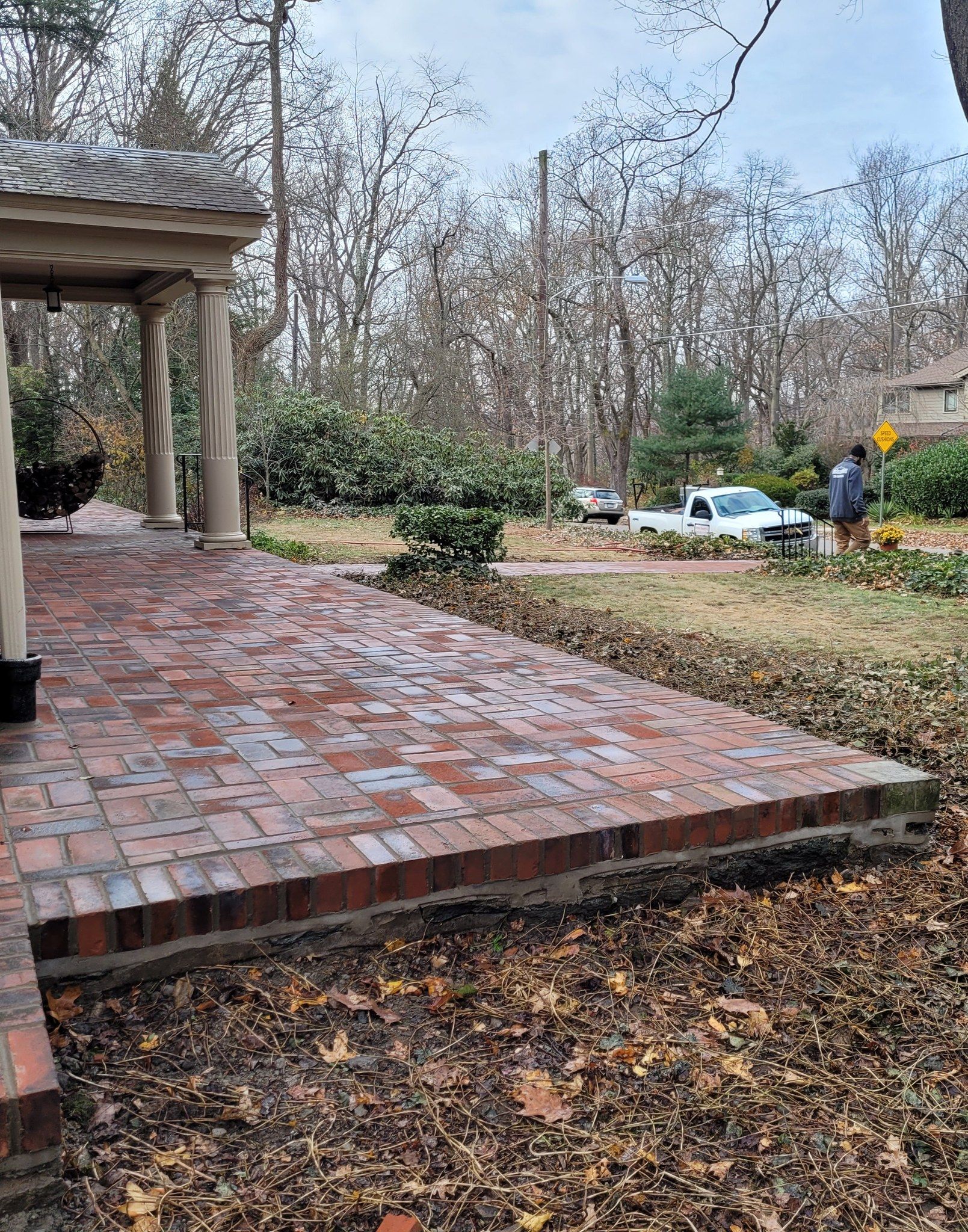 Brick patio with pergola, worker and white truck in the background, surrounded by trees and leaves.