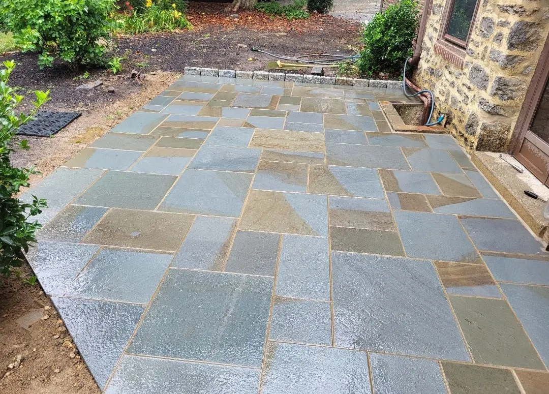 Wet blue-gray stone patio with irregular shapes, next to a stone building, with dirt and plants nearby.
