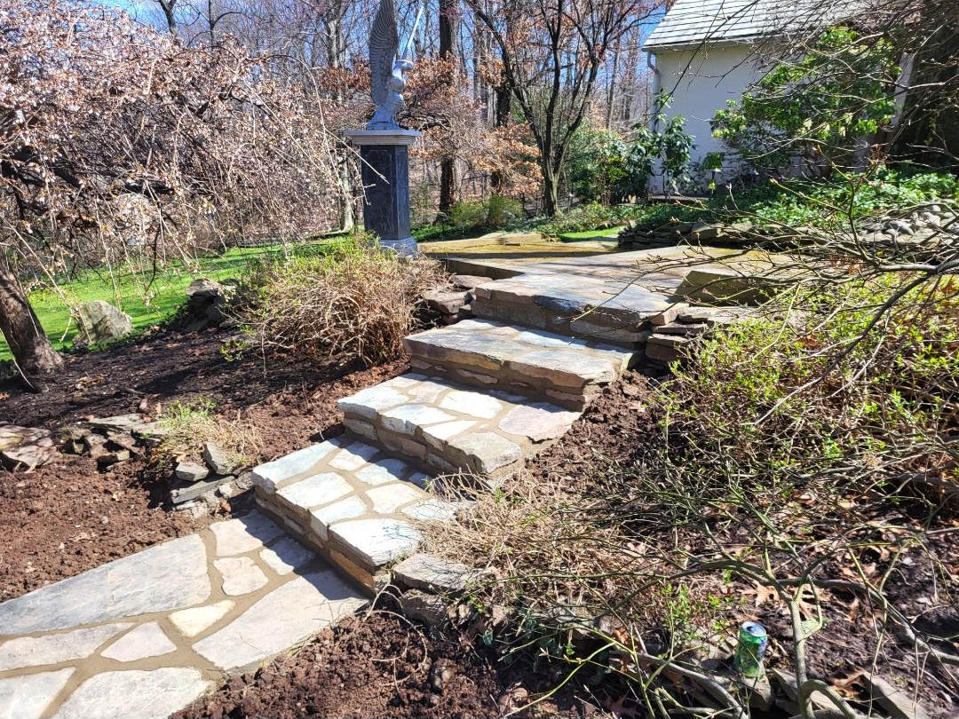Stone steps and pathway lead uphill to a statue and small white building in a wooded garden.