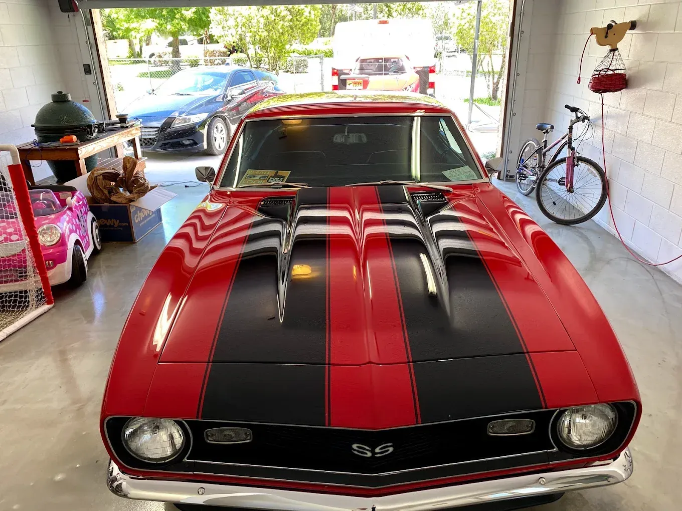 Red classic car with black stripes in a garage, a blue car and a bicycle visible.