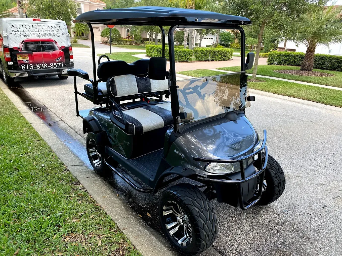 Dark gray golf cart parked on a residential street, next to a green lawn and a service truck.