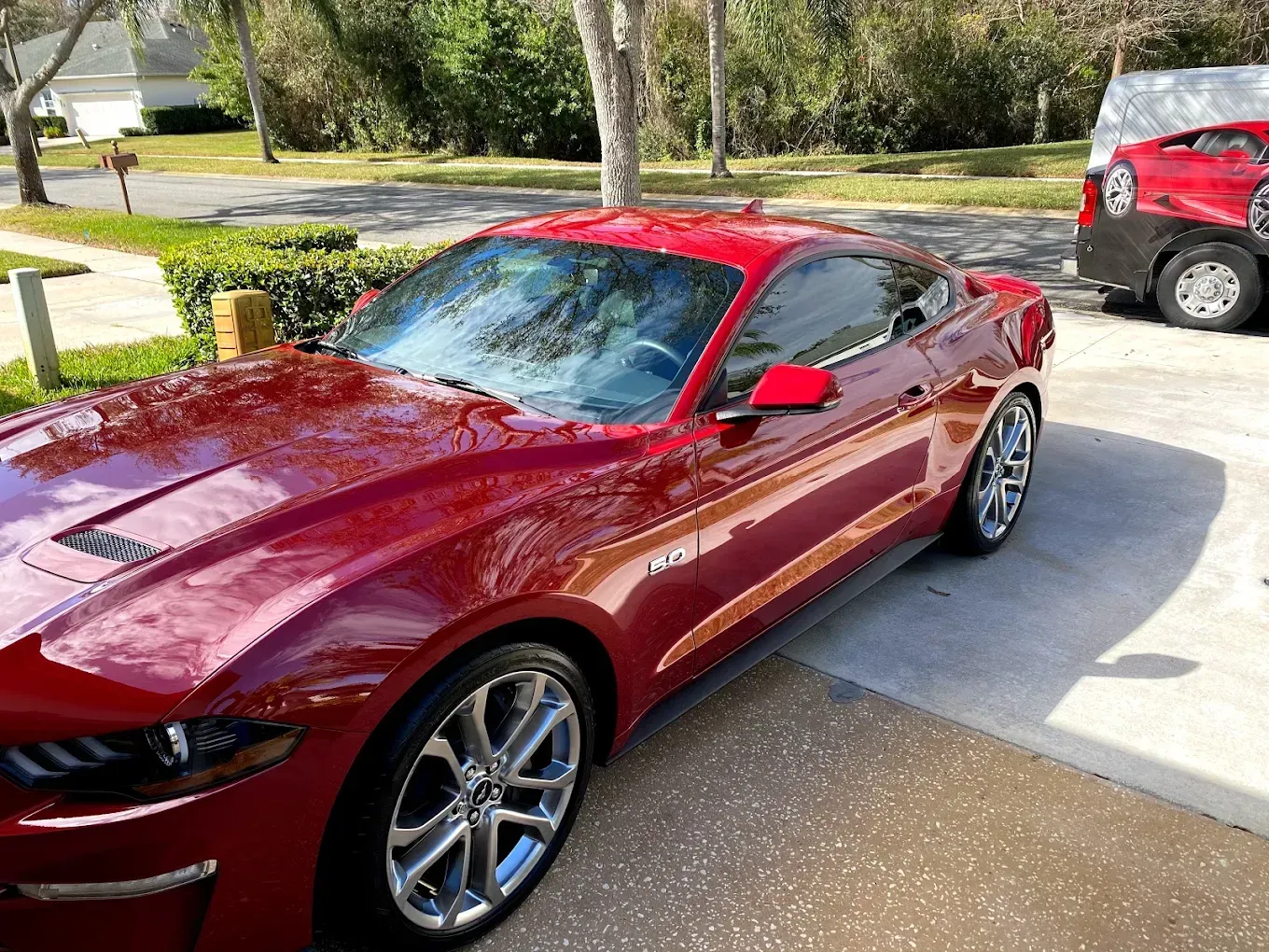 Red Ford Mustang parked on a driveway, reflecting sunlight, with a glimpse of another car in the background.