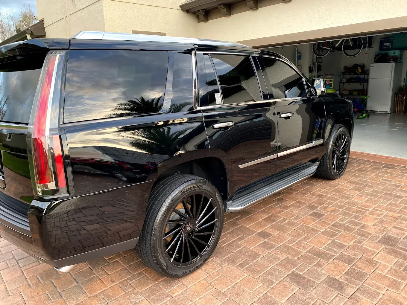 Black Cadillac Escalade parked on a brick driveway, with tinted windows and black rims, in front of a garage.