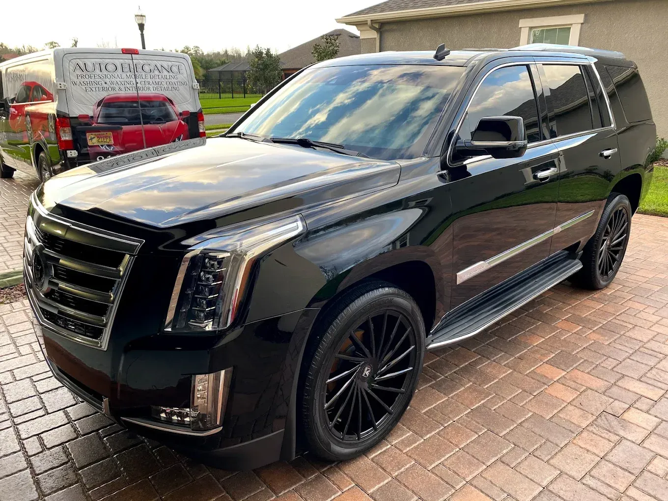 Black Cadillac Escalade parked on a brick driveway; detail of a commercial vehicle in the background.
