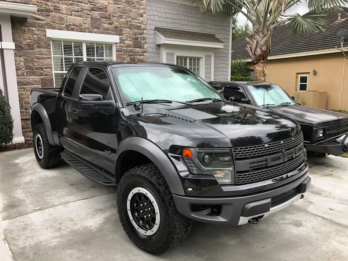 Black Ford Raptor pickup truck parked in front of a house on a paved driveway.