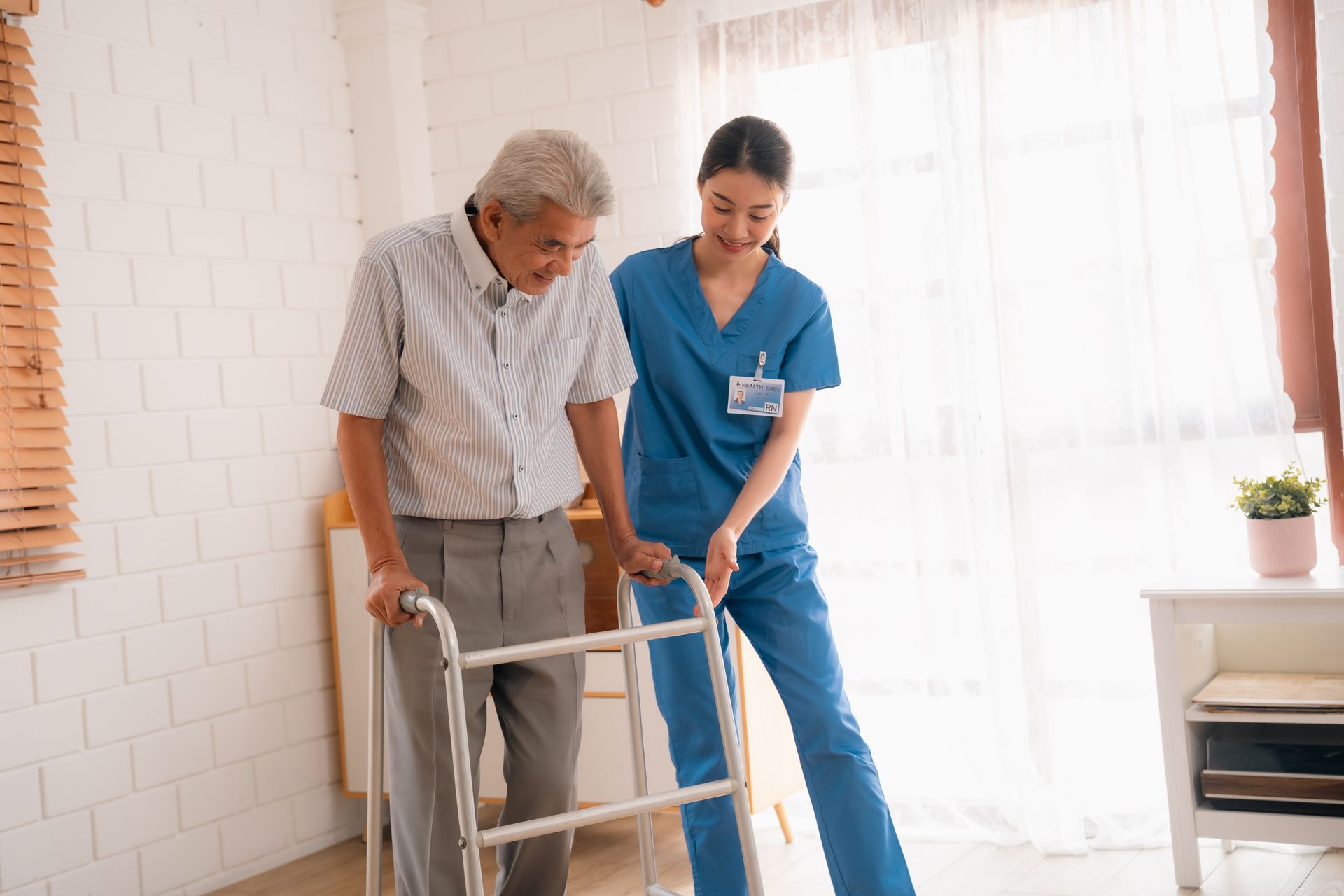 A female nurse in dark blue scrubs assists an older gentleman, inside a house.