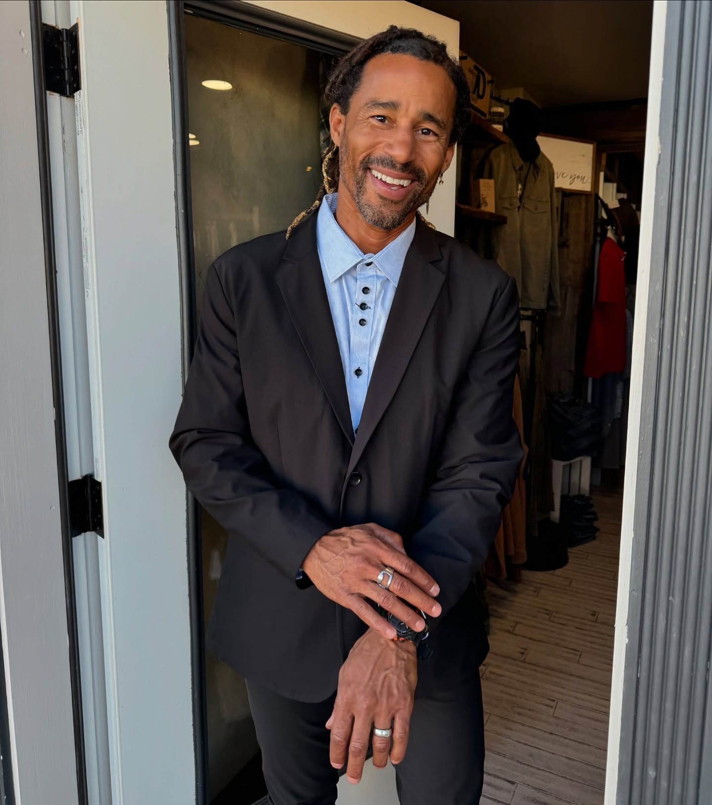 Man in black suit smiles, standing in doorway of a shop.