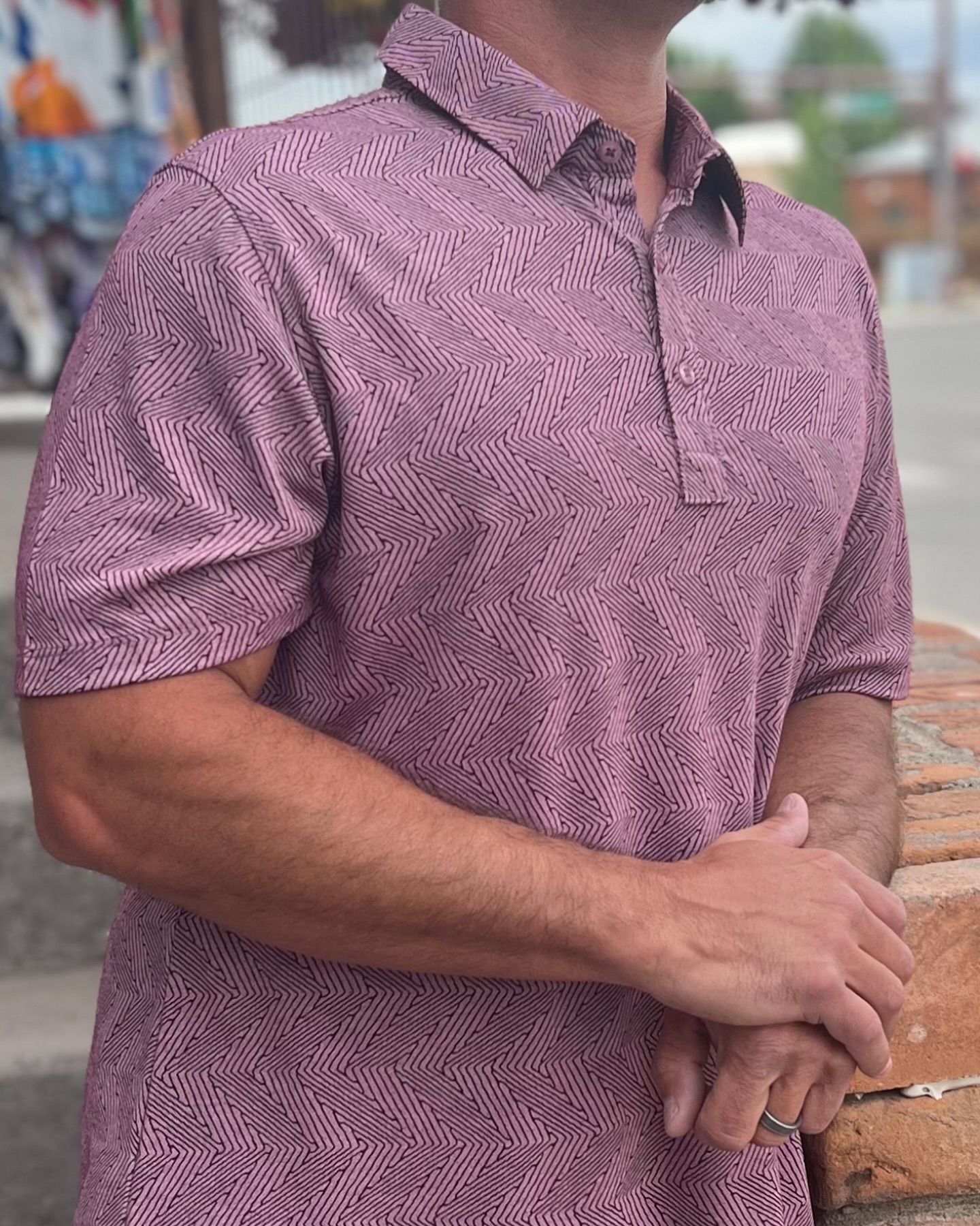 Man in patterned mauve polo shirt, standing outdoors, arm resting on a brick structure.