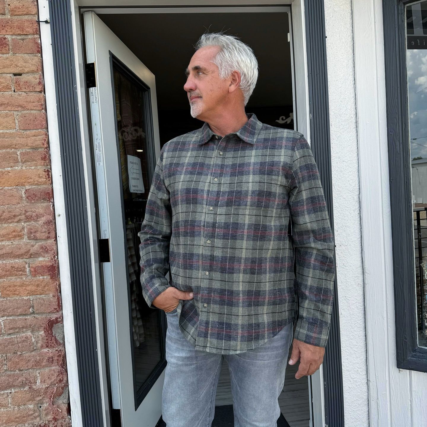 Man in plaid shirt and jeans standing in doorway, looking right, hand in pocket.
