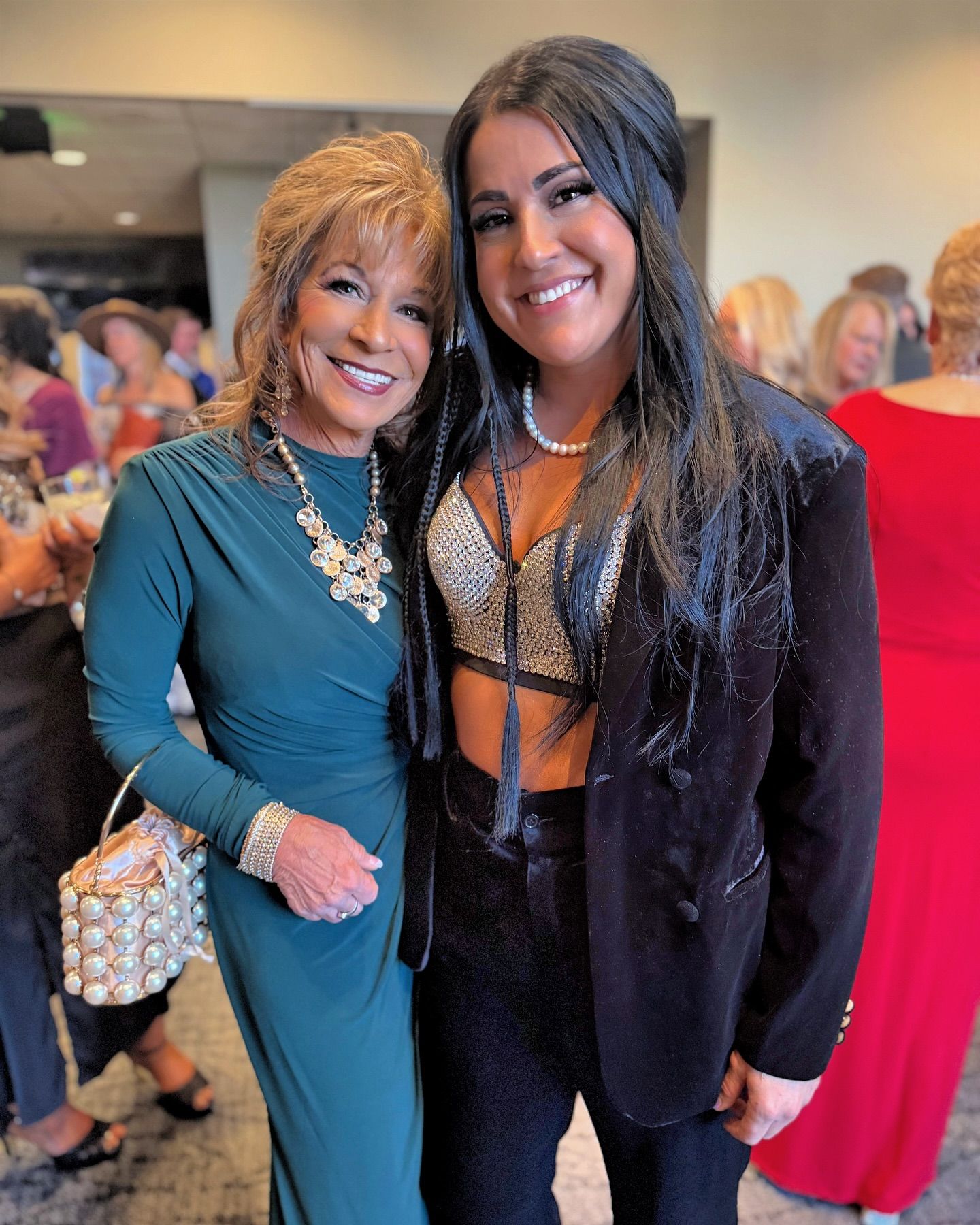 Two women smiling, posing for photo. One in green dress, one in crop top and suit. Indoors, event setting.