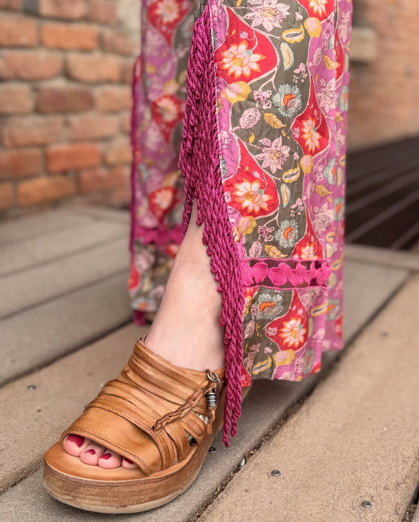 Woman's foot in a brown sandal peeks from floral pants with a pink fringe trim. Outdoors, near brick.