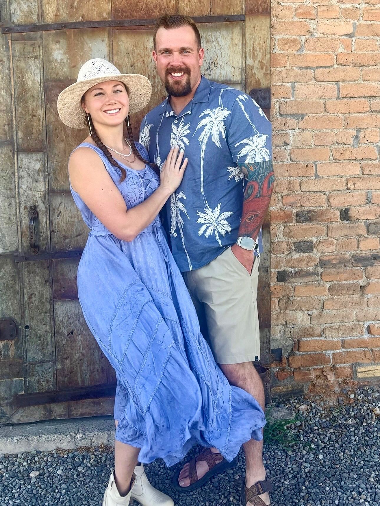 Couple smiling, posing in front of a rustic door and brick wall. Woman wears a blue dress and straw hat; man, a blue shirt with shorts.