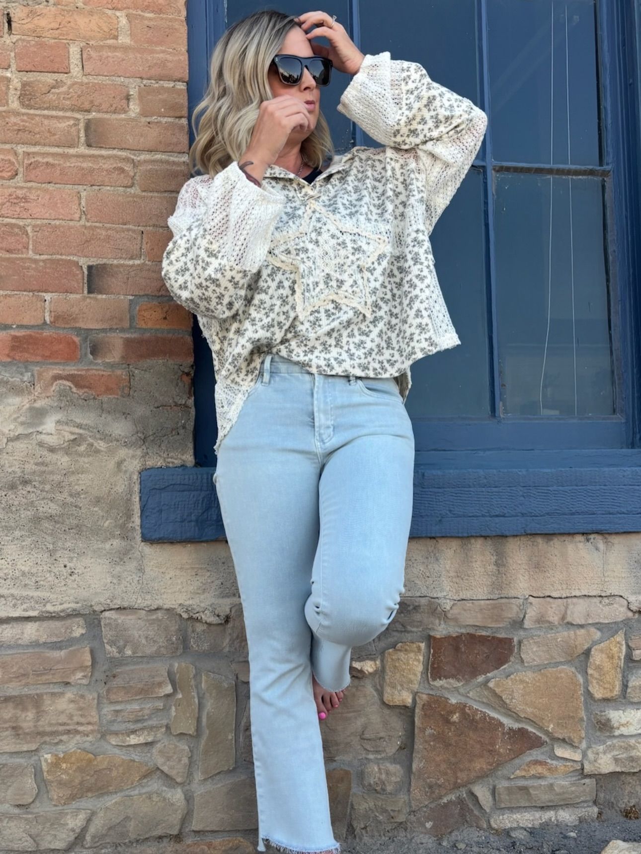 Woman in light-colored floral top and light blue jeans leaning against a brick wall and blue window.