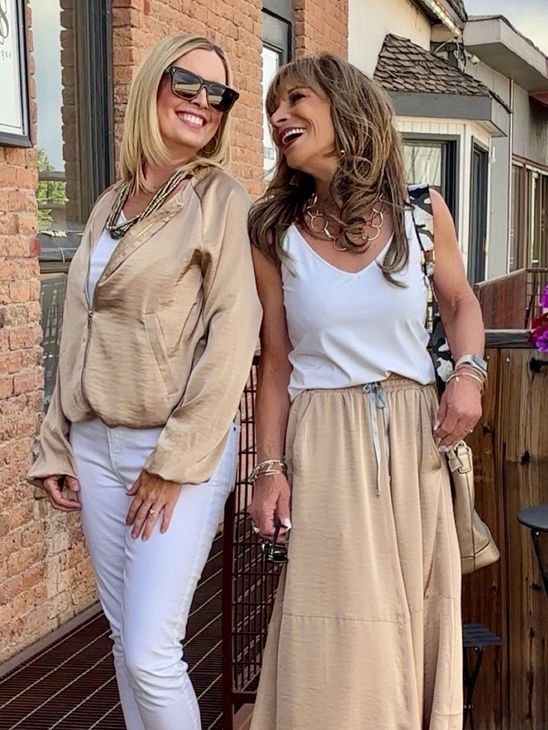 Two women smiling outdoors; one in white, tan jacket, sunglasses, the other in white, tan skirt, necklace.
