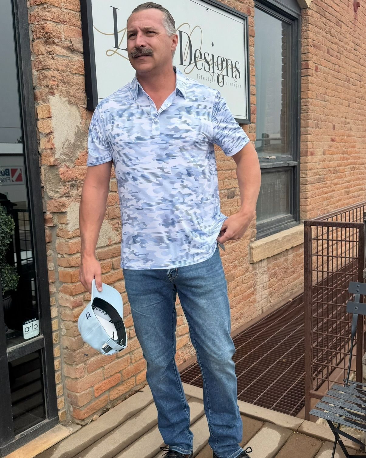 Man wearing a blue patterned polo shirt and jeans, holding a hat, standing near a brick building.