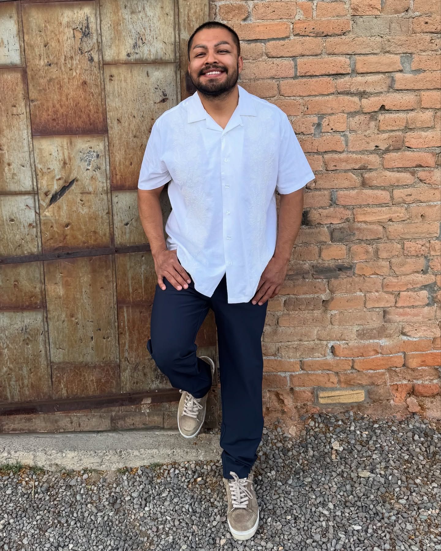 Man in white shirt and navy pants leans against a metal door and brick wall, smiling.