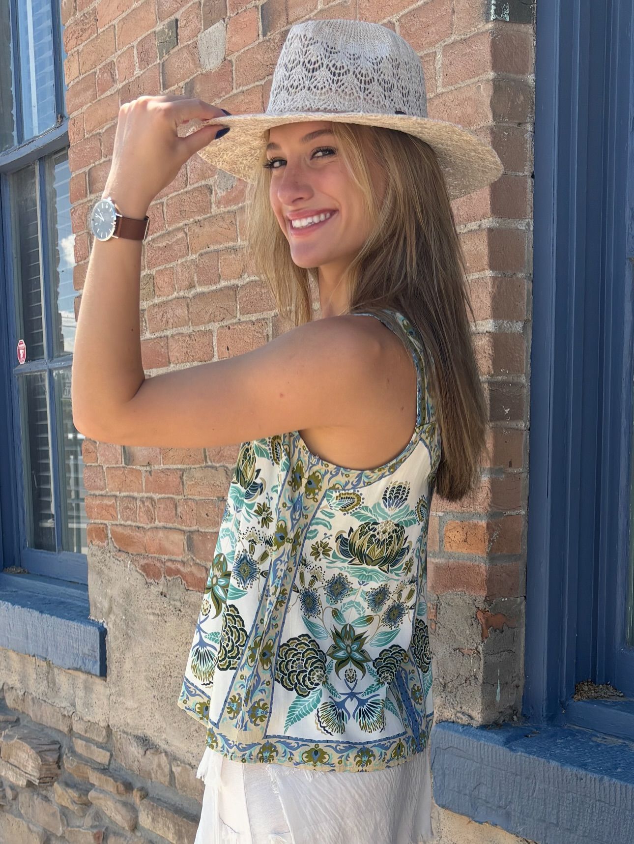 Woman wearing a hat and patterned top smiling by a brick building.