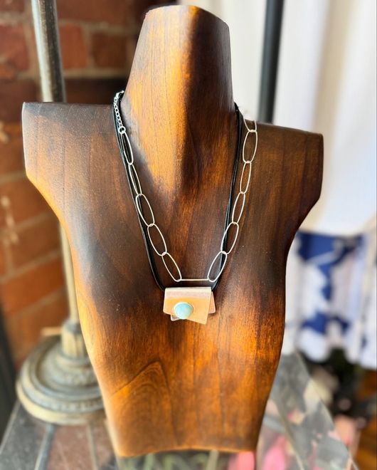 Wooden bust holding a silver necklace with a rectangular pendant and a light blue stone.