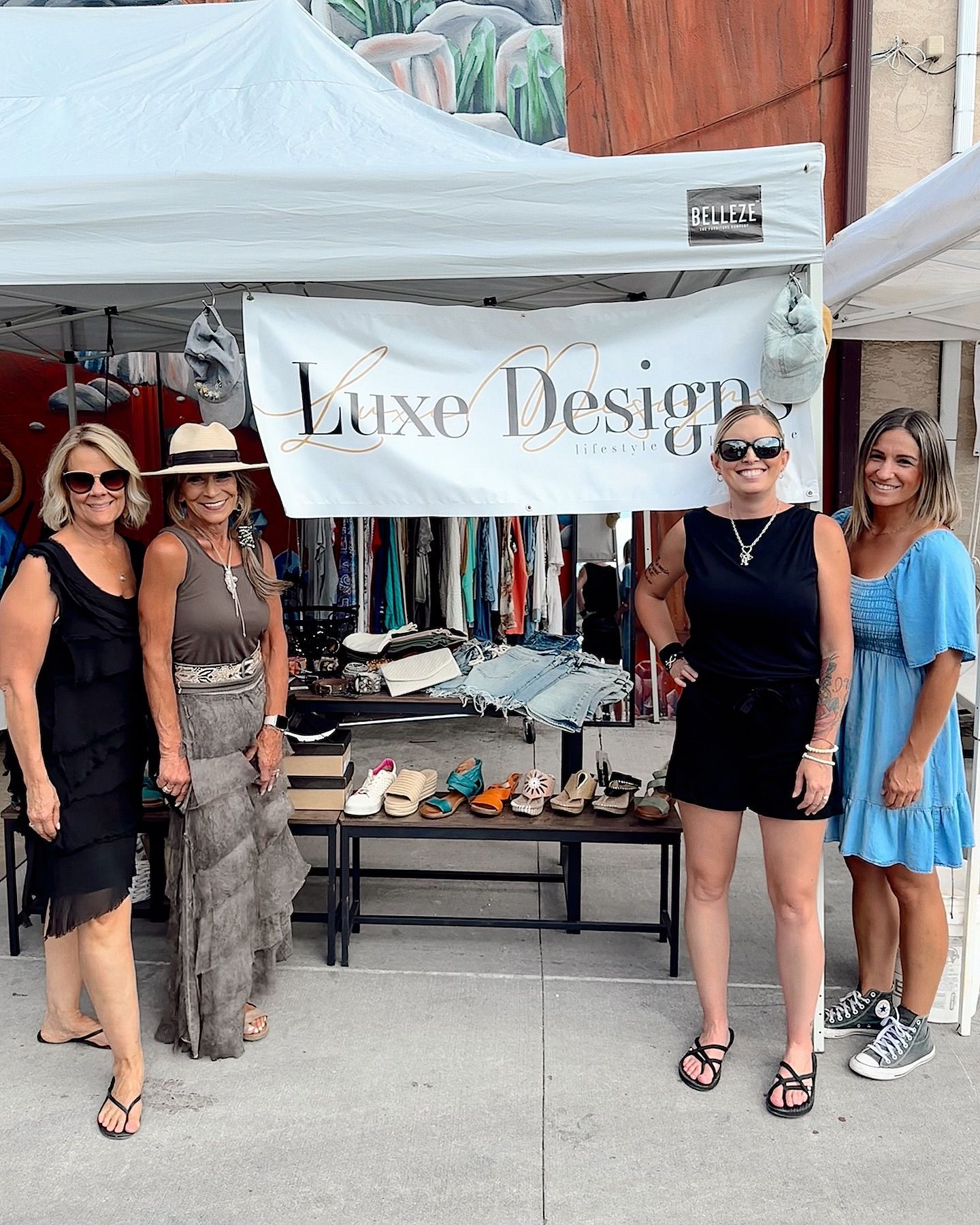 Four women stand in front of a Luxe Design booth. A variety of clothing and shoes are displayed.
