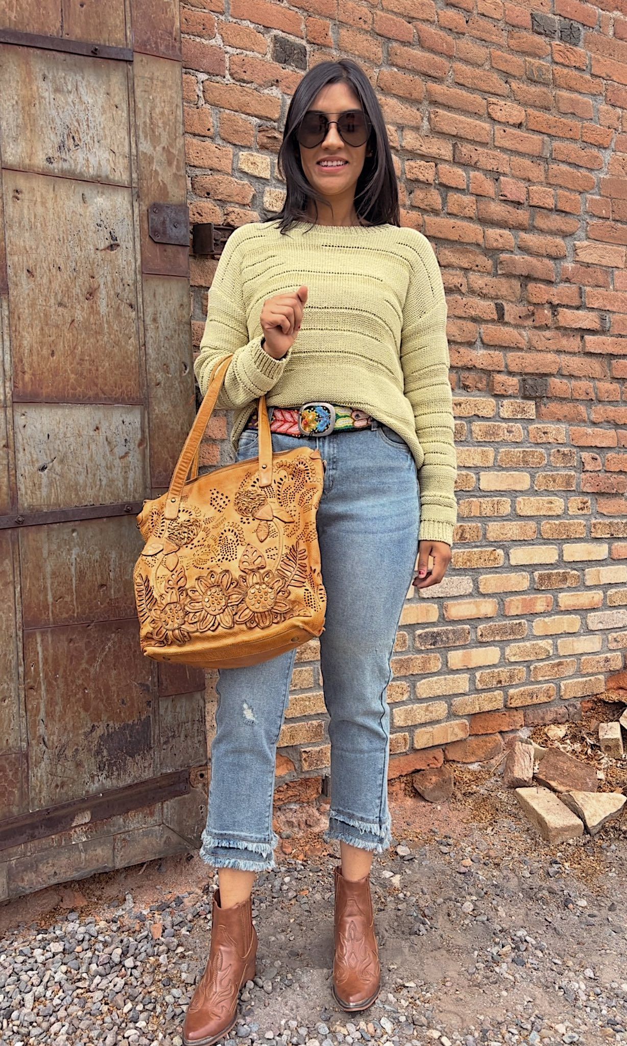 Woman in light green sweater, blue jeans, and brown boots stands by a brick wall, holding a tan tote bag.