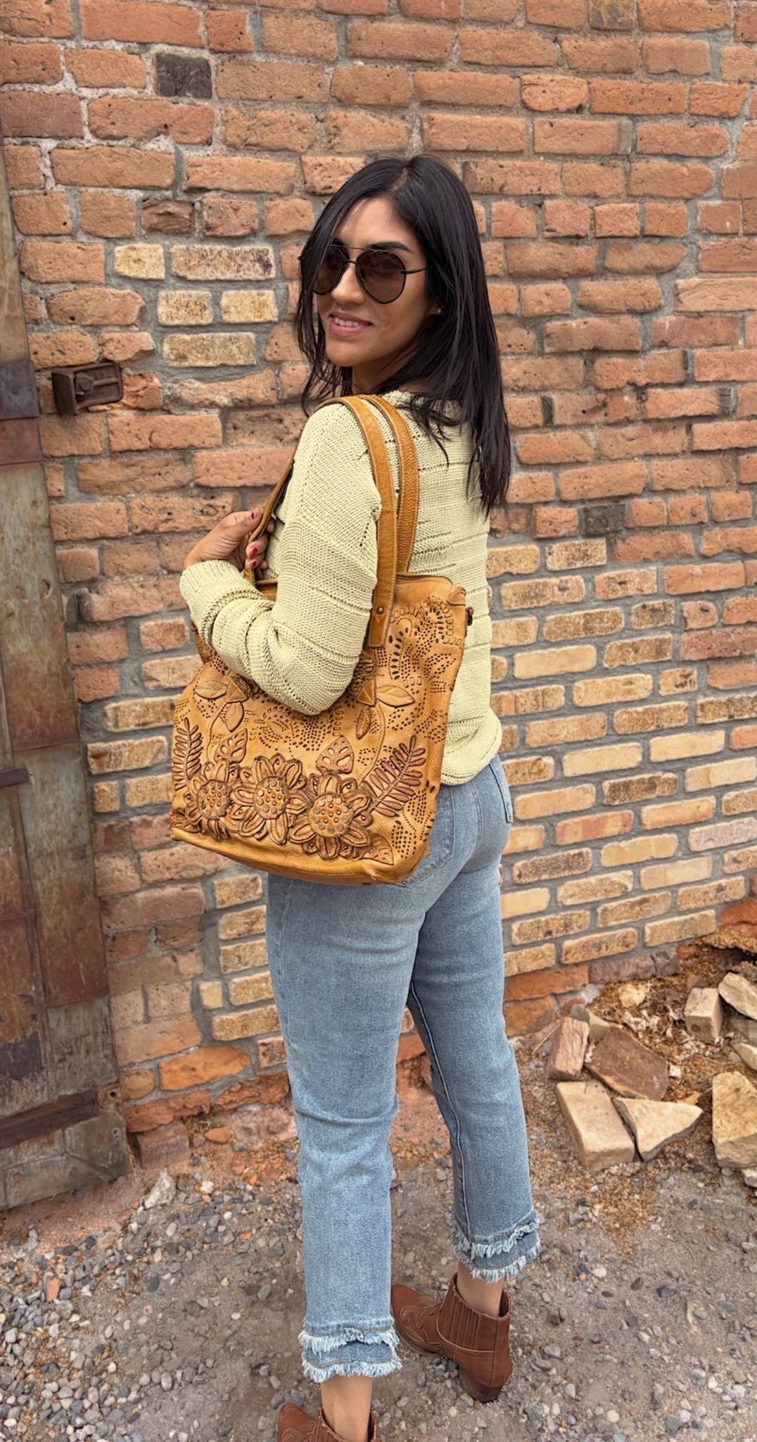Person in light green sweater and jeans, brown bag, sunglasses, against a brick wall.