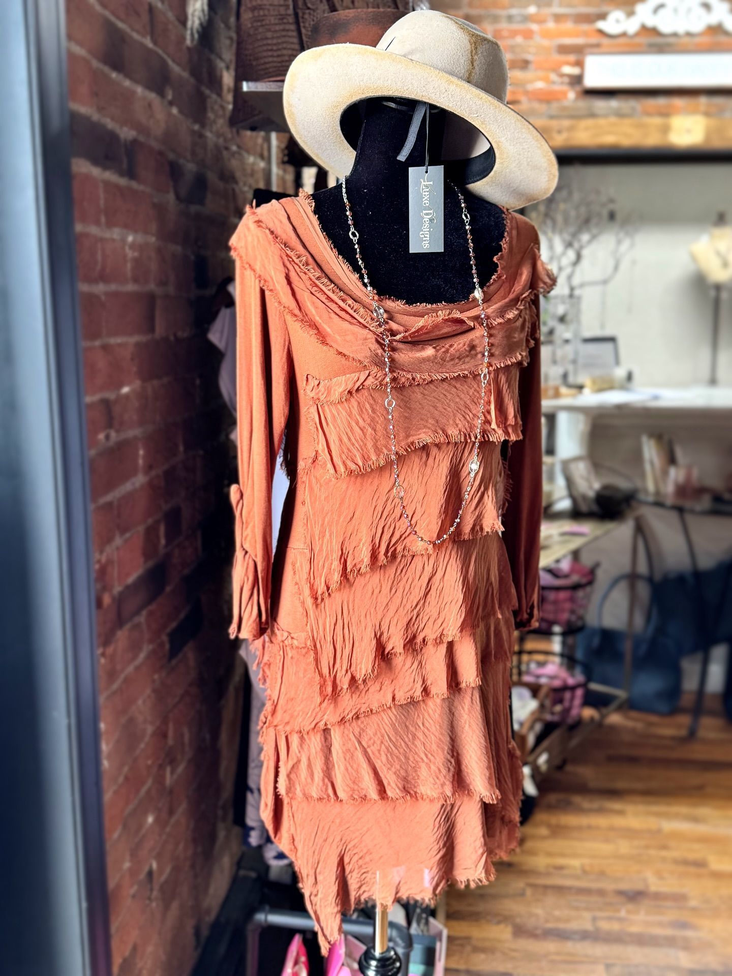 Dress on a mannequin in a shop window. Rust-colored tiered dress with a straw hat and necklace. Brick wall background.