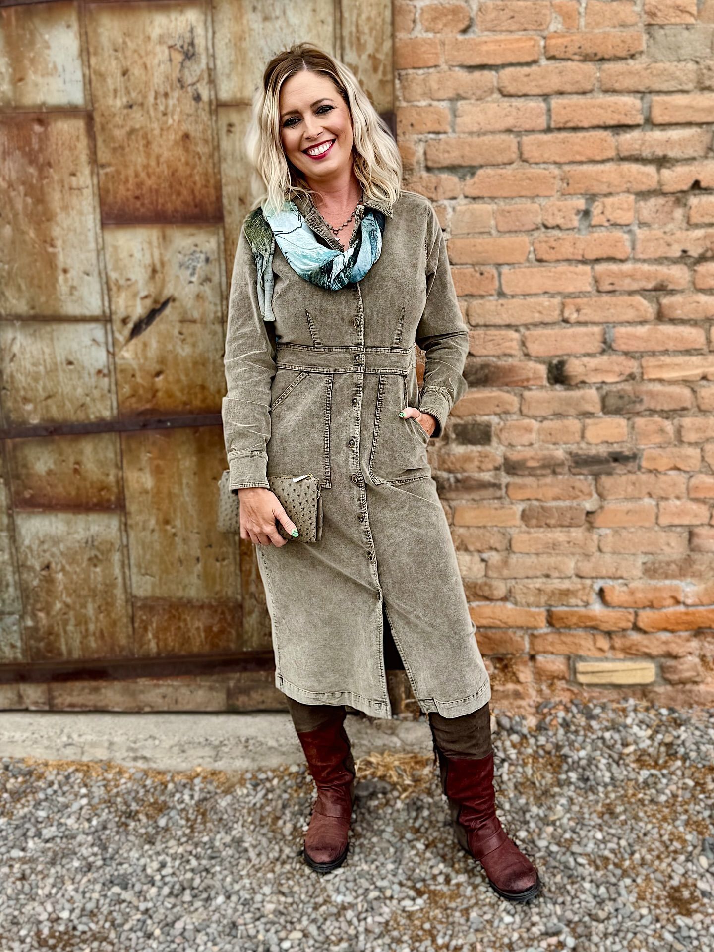 Woman in olive green coat and boots stands in front of a brick wall and rustic door.