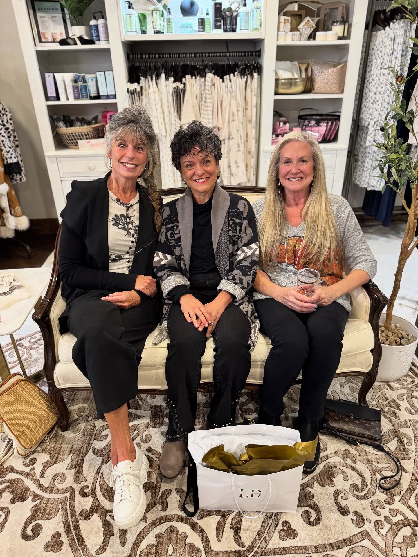 Three women seated on a sofa in a boutique setting; smiles, casual attire, and shopping bags are present.