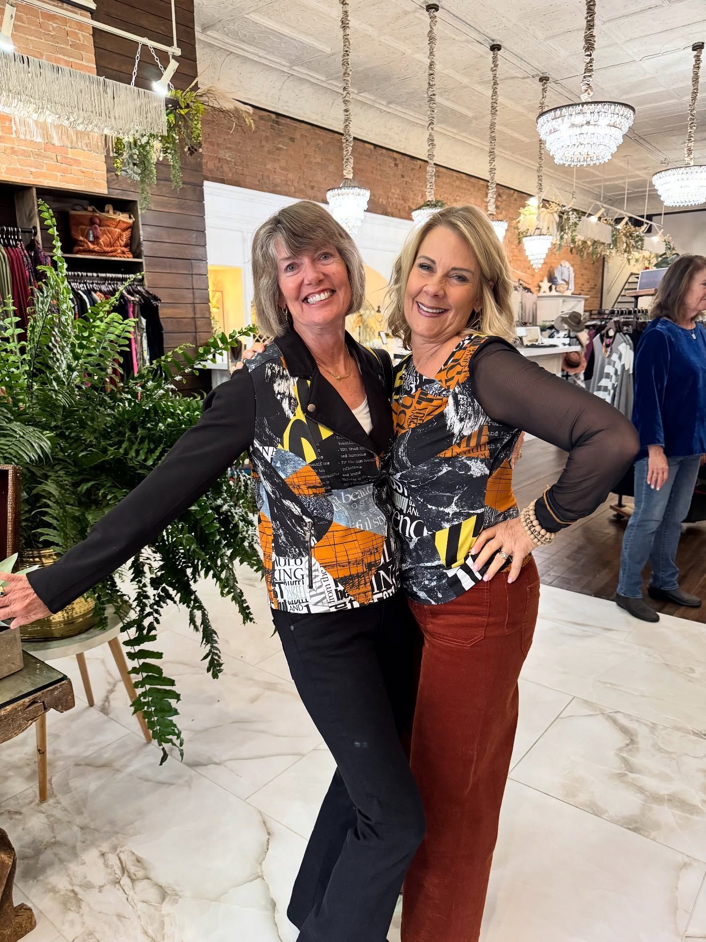 Two women smiling, posing in a boutique; wearing patterned tops, black pants, and corduroy pants.