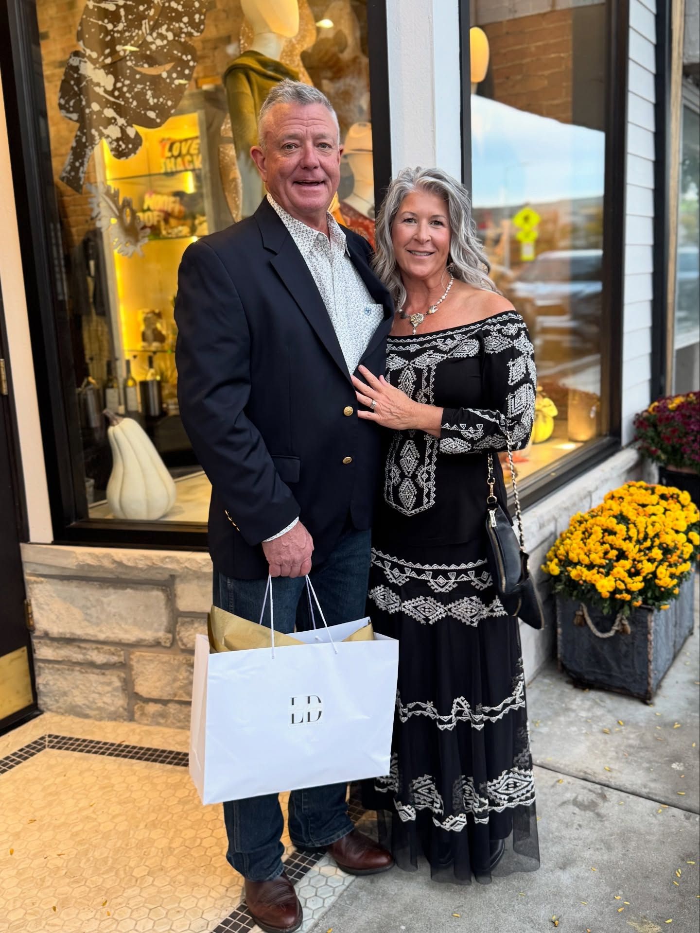 Couple standing in front of shop window, woman in black dress, man in blazer, holding shopping bag.