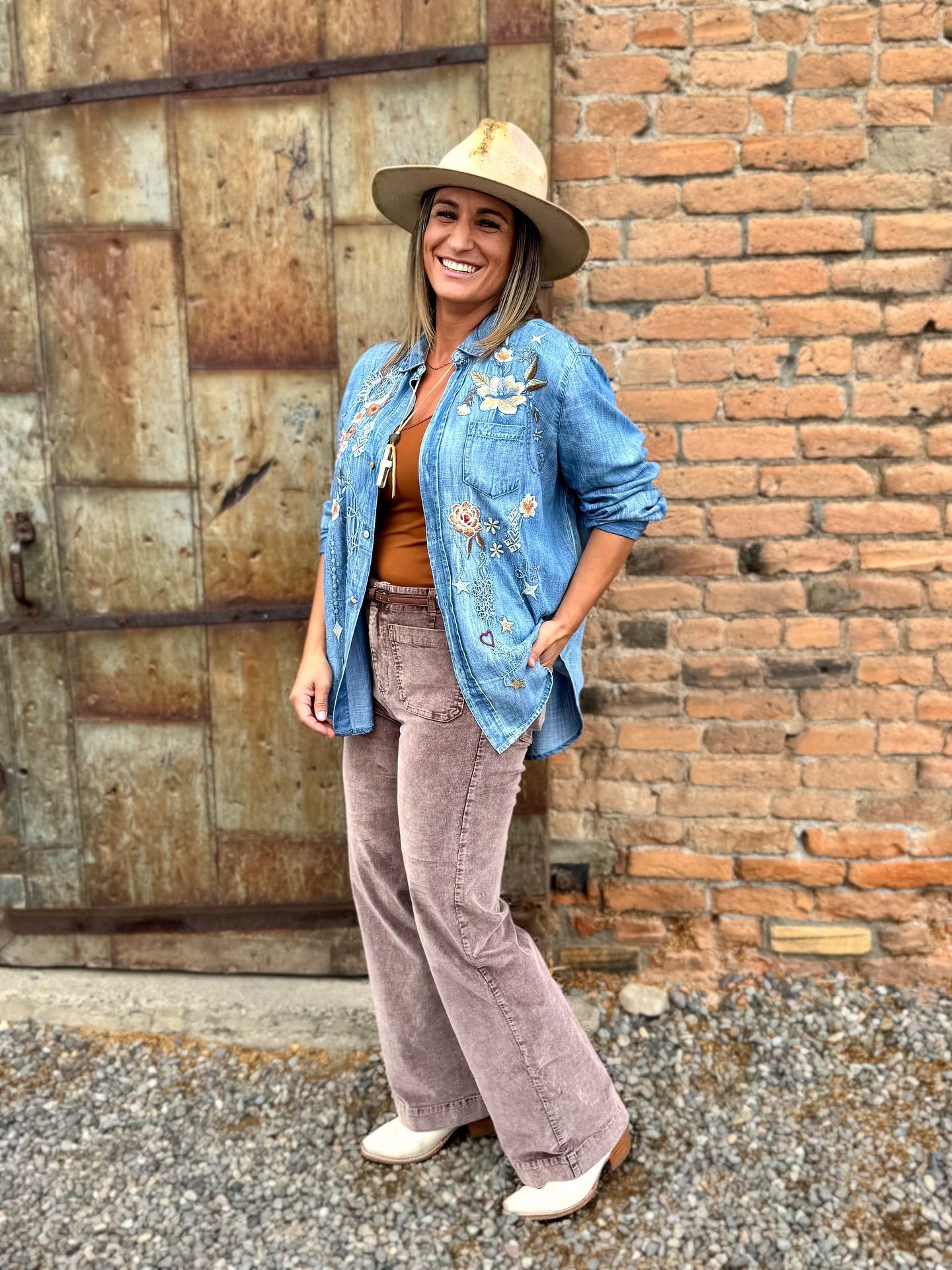 Woman in denim jacket, hat, and brown pants, smiles by brick wall and metal door.