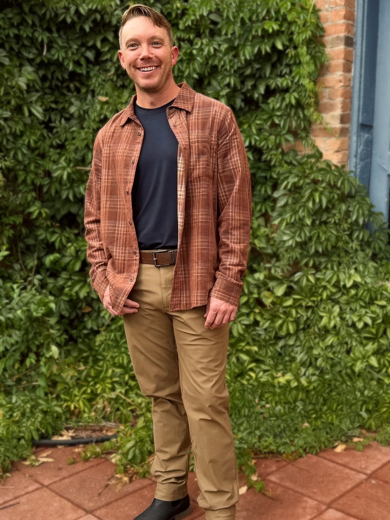 Man in brown plaid shirt and khaki pants, standing in front of ivy-covered wall.