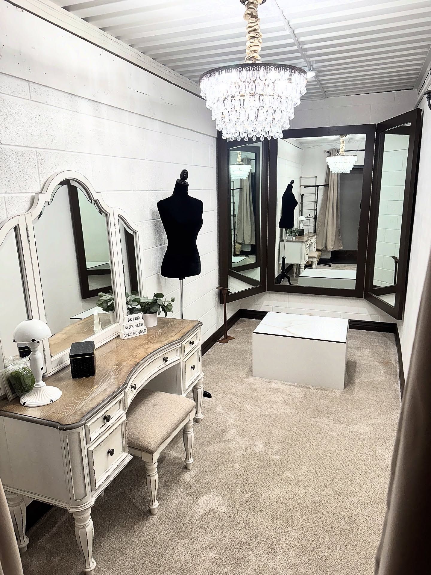 Vanity area with a chandelier, three mirrors, a dress form, and a white bench on a beige carpet.