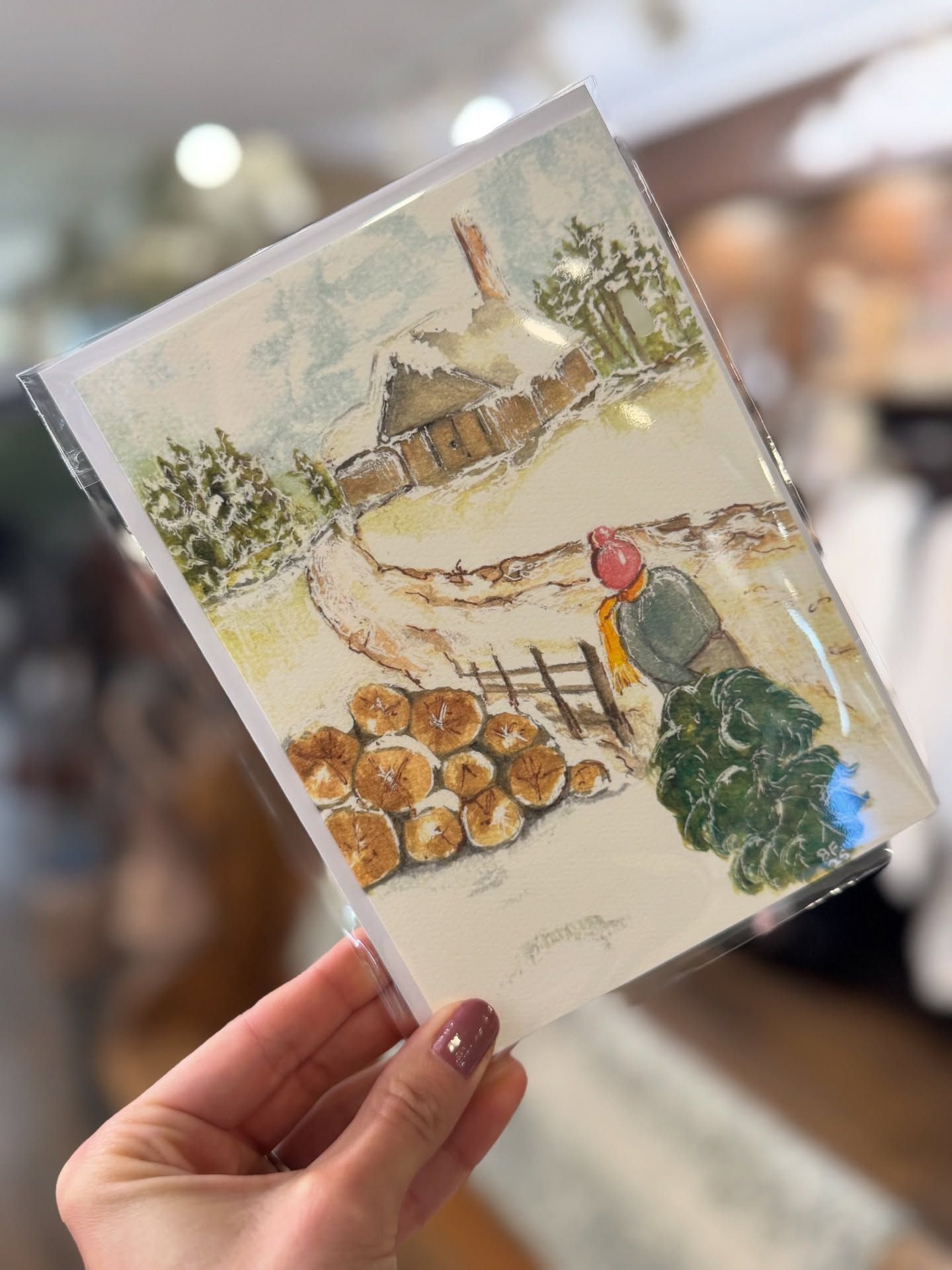 Watercolor painting of a snowy scene. A person carries a green tree past a log pile and cottage.
