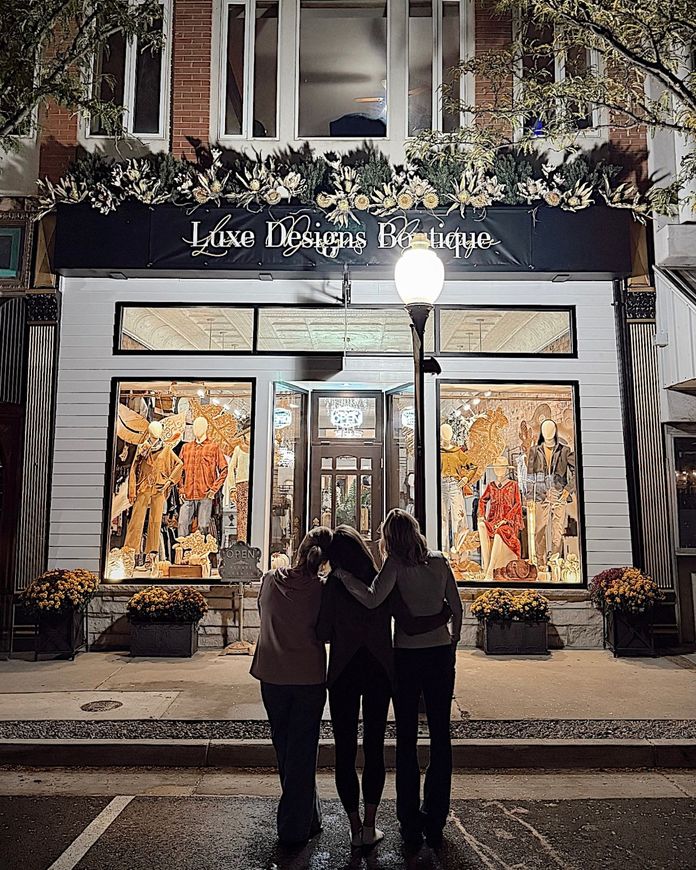 Three people embrace in front of a storefront at night, decorated with lights and floral arrangements.
