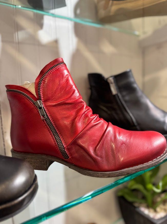 Red leather ankle boot with side zipper, displayed on a shelf. Another black boot is visible in the background.
