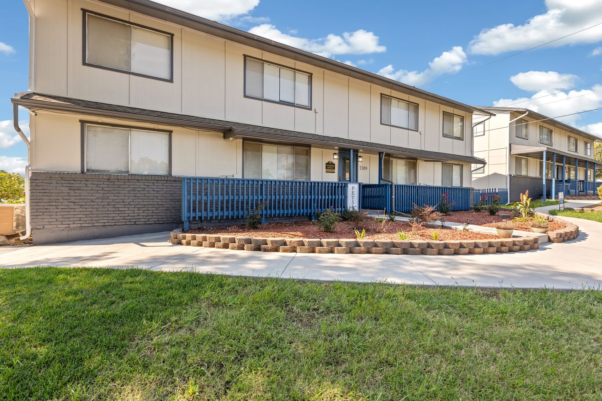 Blue Sky Apartments, tan building with blue trim, sidewalk, green bushes, trees, and grass.