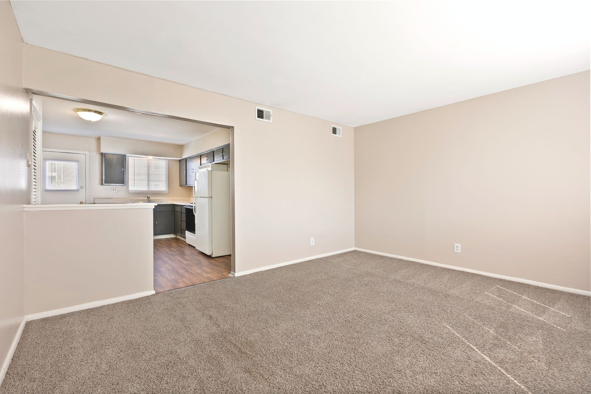 Interior view of a living room with grey carpet, kitchen visible through an opening.