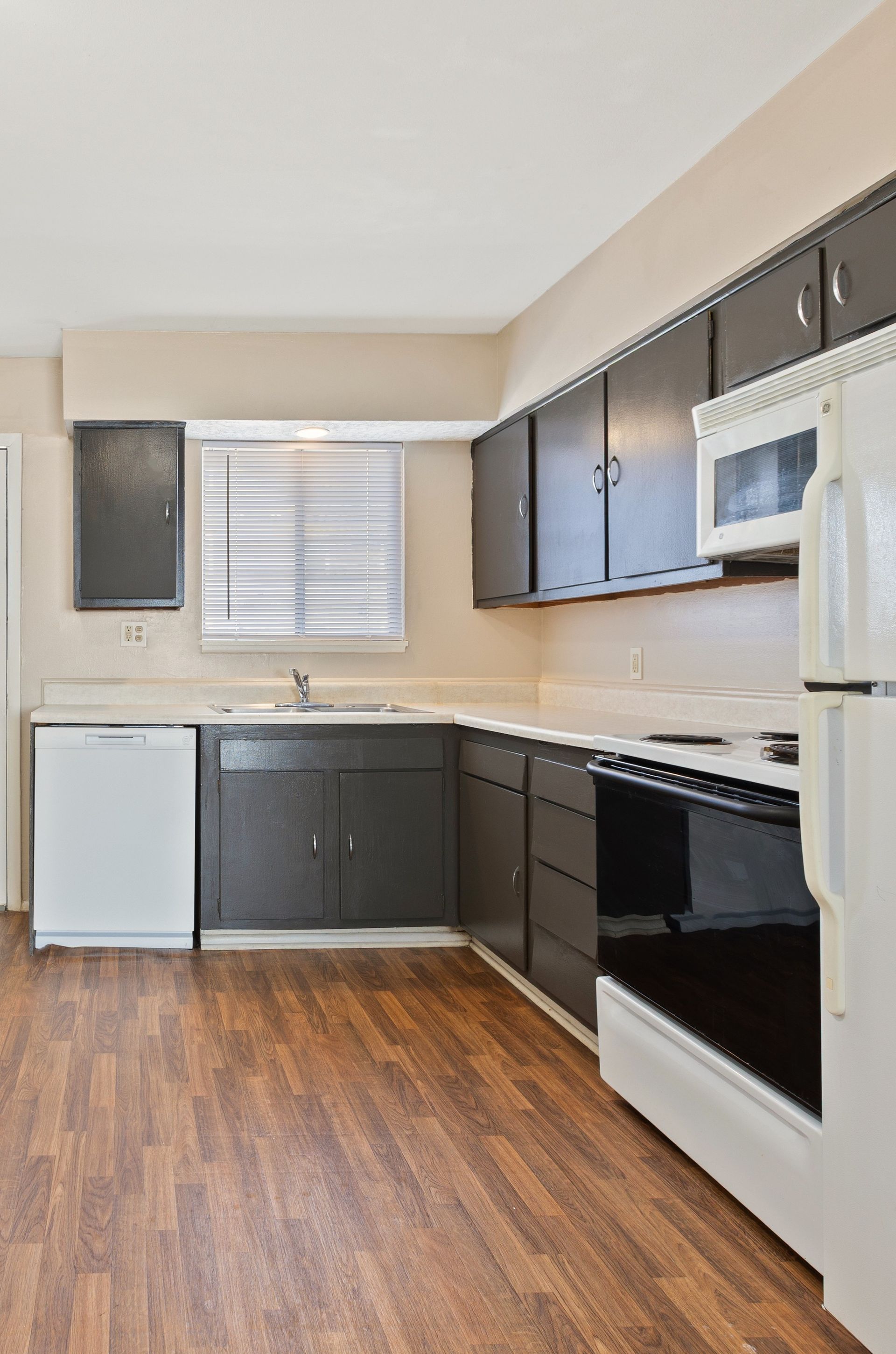 Spacious kitchen with dark cabinets, white appliances, and wood-look flooring. Window with blinds.