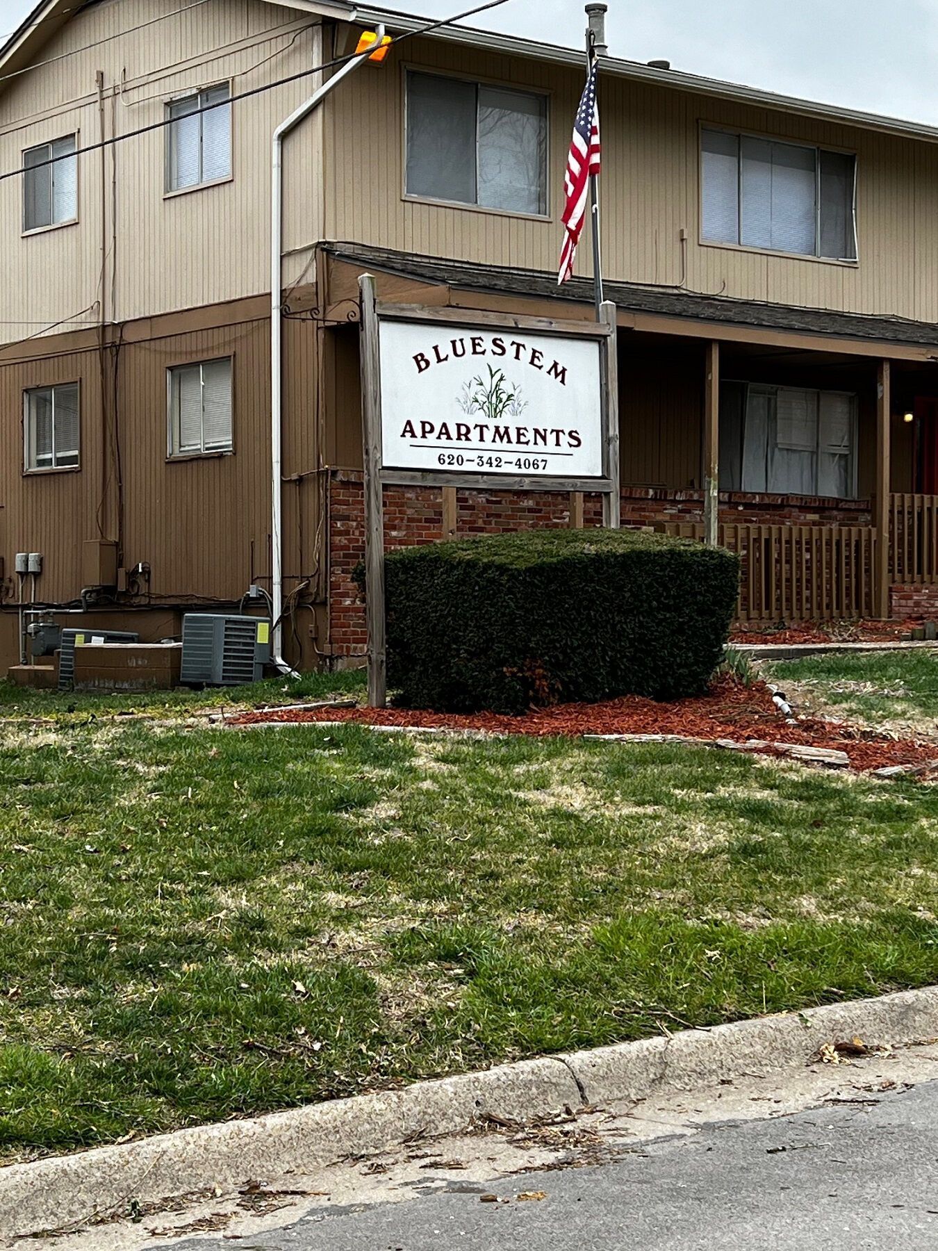 Blue Sky Apartments sign with US flag, brown building, green bushes, and grass.