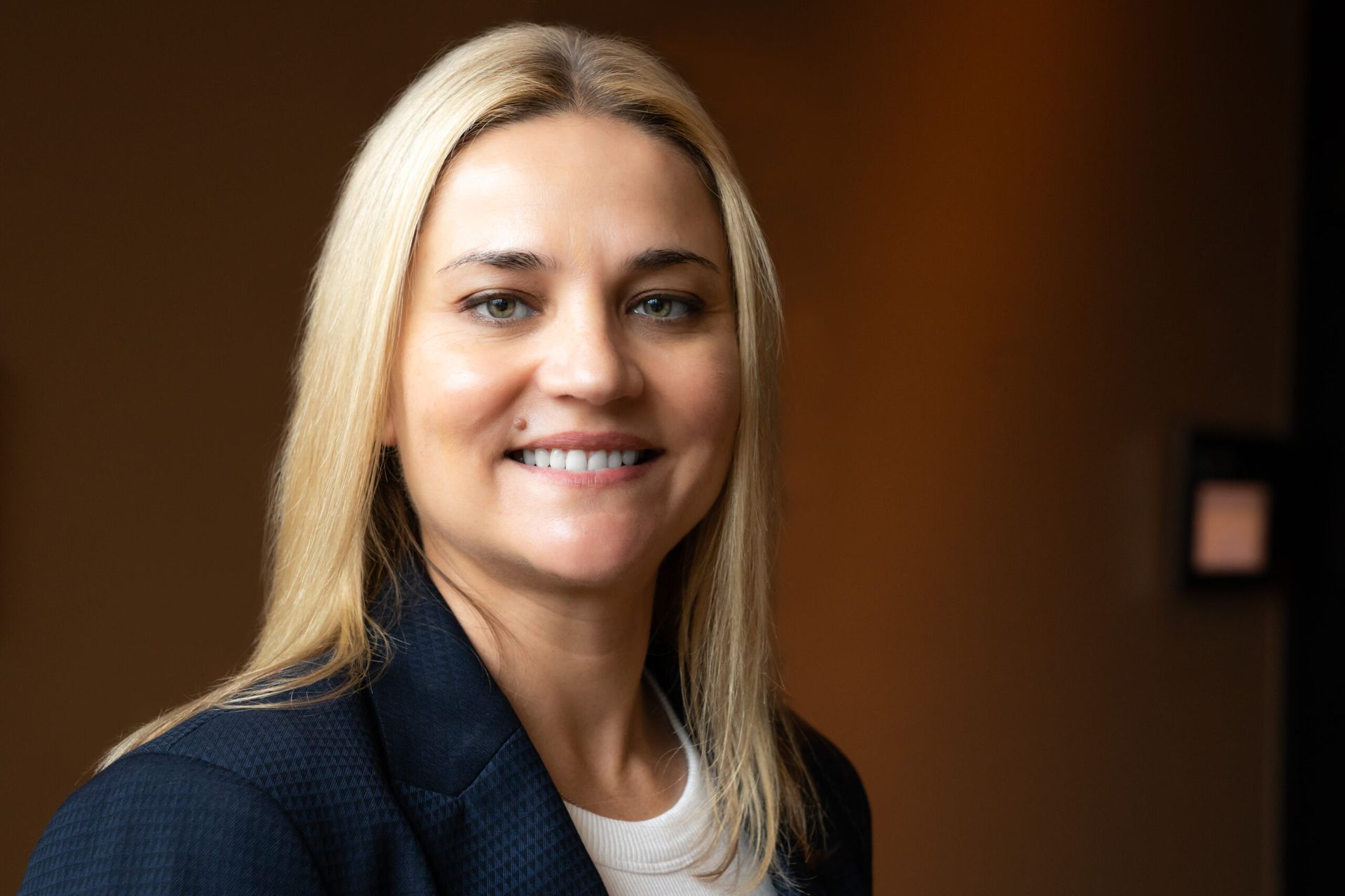 Blonde woman with a slight smile wearing a dark blazer and white shirt, in a room with a brown wall.