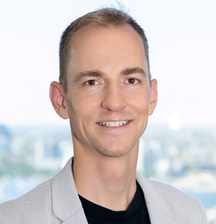 Man with short brown hair, smiling, wearing a light grey jacket. City background.